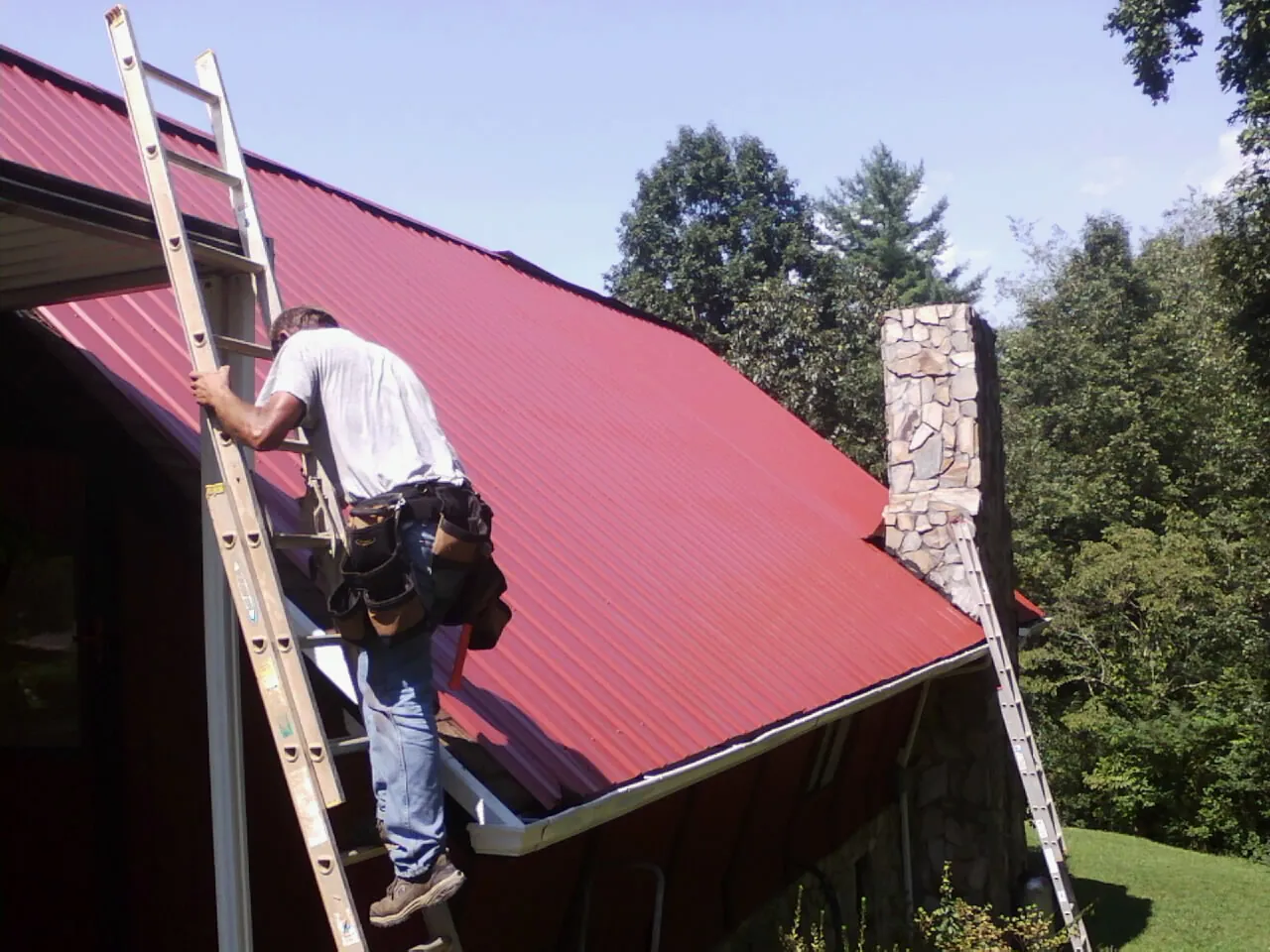 Roofer on a ladder beside a red metal roof and stone chimney — Soffit & Fascia Repair by Roofing Brothers in Lakemore